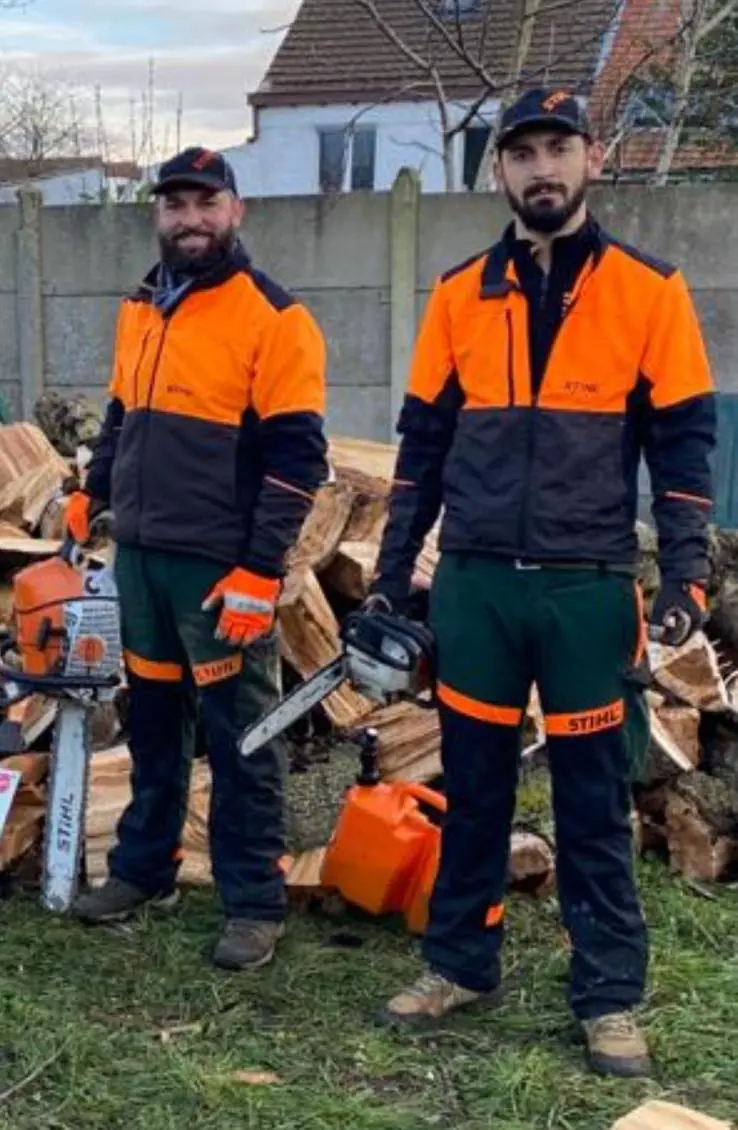 Jean et Matthieu en tenue d’élagueur, tronçonneuse à la main, devant un tas de bois